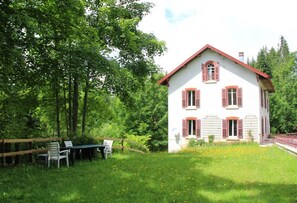 Une maison blanche aux volets rouges, entourée d’une clôture en bois et d’un espace verdoyant avec une table et des chaises.