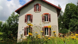 A two-story house with red shutters and a white facade, surrounded by a garden with yellow flowers. - LE GRAND TETRAS in a charming property in the heart of the Blue Spring area (Montperreux)