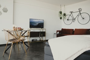 A modern living room with a bed, a dining area with a table and chairs, a TV mounted on the wall, and a bicycle hanging on the wall.
