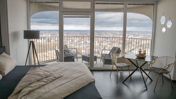 Quarto moderno com uma cama grande, uma escrivaninha e uma cadeira. O quarto possui uma varanda com vista para a cidade.