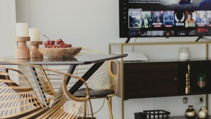 Uma área de refeições moderna com mesa de vidro, cadeiras de vime e uma televisão de tela plana na parede exibindo um filme.