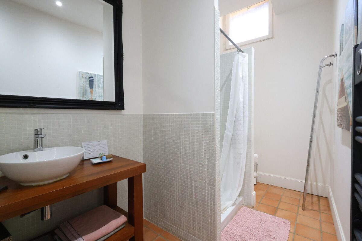 Un baño con lavamanos blanco, tocador de madera, paredes con azulejos, ducha con puerta de vidrio y una ventana pequeña.