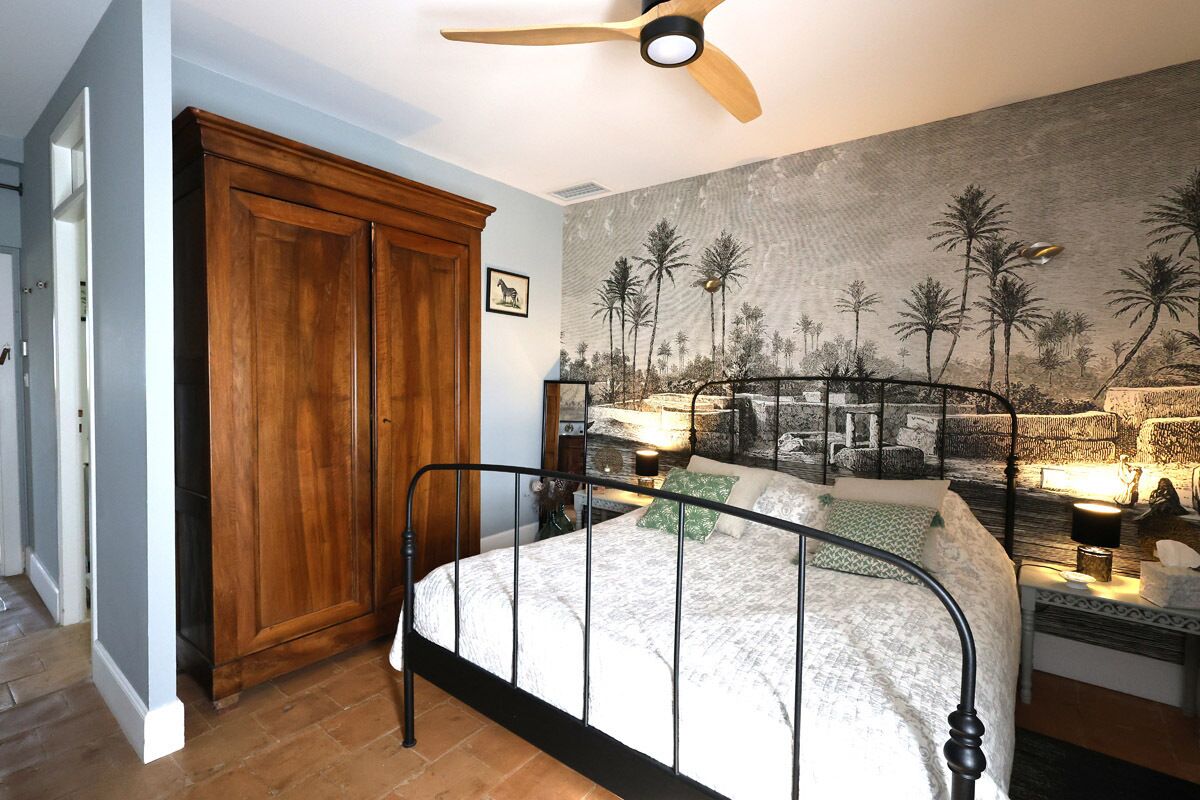 A bedroom with a bed, a wooden wardrobe, a ceiling fan, and a mural on the wall.