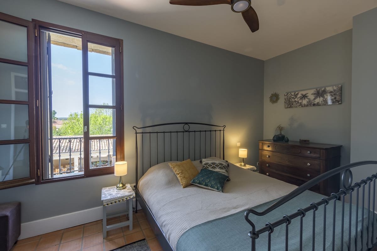 Quarto com cama, mesas de cabeceira, cômoda, ventilador de teto e janela com vista.