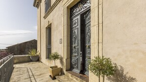 A building with ornate black doors, a balcony with potted plants, and a stone wall in the background. - Maison Dix (Neffies)