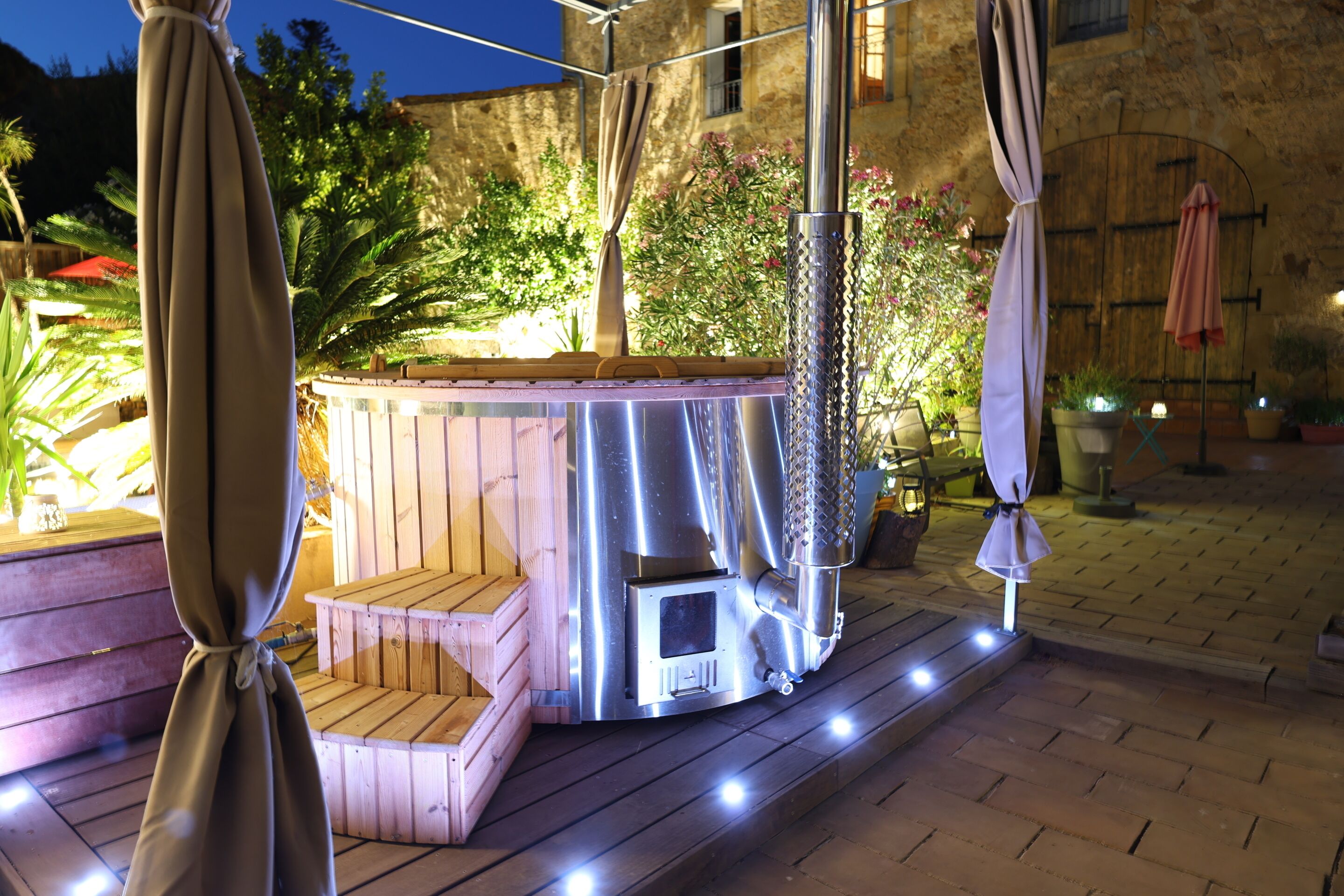Een jacuzzi met een houten bank, omringd door potplanten en buitenverlichting.
