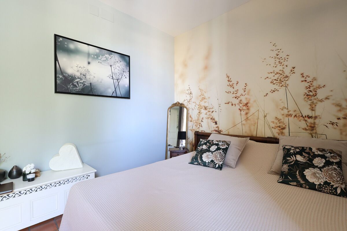 A bedroom with a bed, a bedside table, a mirror, and a wall with a floral mural.