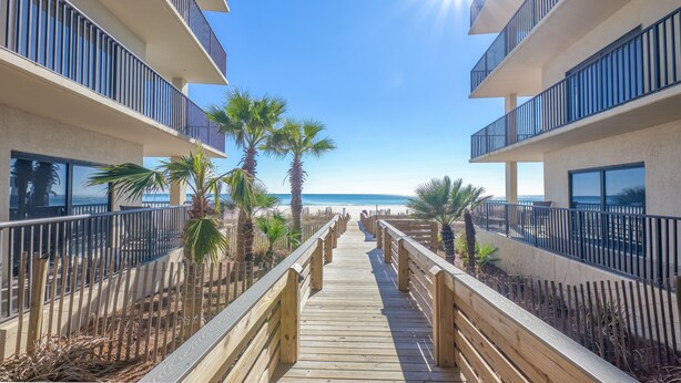 A wooden boardwalk leading to a beach, flanked by buildings with balconies and palm trees. - RELAX AT THE  BEACH IN THIS GORGEOUS GULF FRONT CONDO! THE PALMS #914 (Orange Beach)