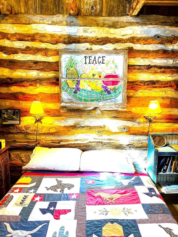 A rustic cabin interior with a bed, a framed artwork of fruits and the word 'PEACE,' and two wall-mounted lamps.