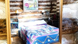 A rustic room with a wooden ceiling, a bed with a colorful quilt, a wall-mounted picture, and a small shelf with books.