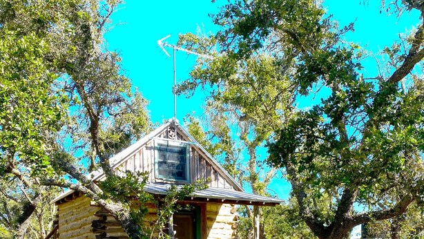A rustic log cabin with a wooden porch and a picket fence, surrounded by lush greenery and trees.