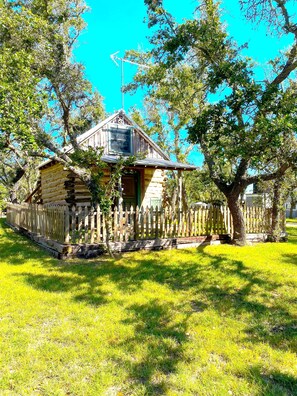 A rustic log cabin with a wooden porch and a picket fence, surrounded by lush greenery and trees.