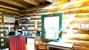 A rustic cabin interior with a wooden dining table, chairs, a small kitchenette, and a 'No Smoking' sign.