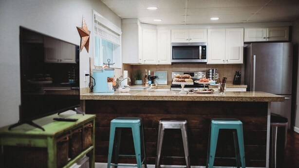 A kitchen with white cabinets, a stainless steel refrigerator, a microwave, and a central island with stools. - Raven’s Nest - Cozy, Northern Lights, Private (North Pole)
