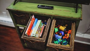 A green wooden table with a remote control on top and two wicker baskets underneath. One basket contains books, and the other has colorful toy parts. - Raven’s Nest - Cozy, Northern Lights, Private (North Pole)