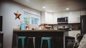 A kitchen with white cabinets, a microwave, a refrigerator, and a bar area with stools. - Raven’s Nest - Cozy, Northern Lights, Private (North Pole)