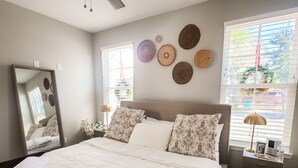 Une chambre à coucher avec un grand lit, un ventilateur de plafond, un miroir, une table de chevet avec une lampe et des œuvres d’art décoratives au mur.