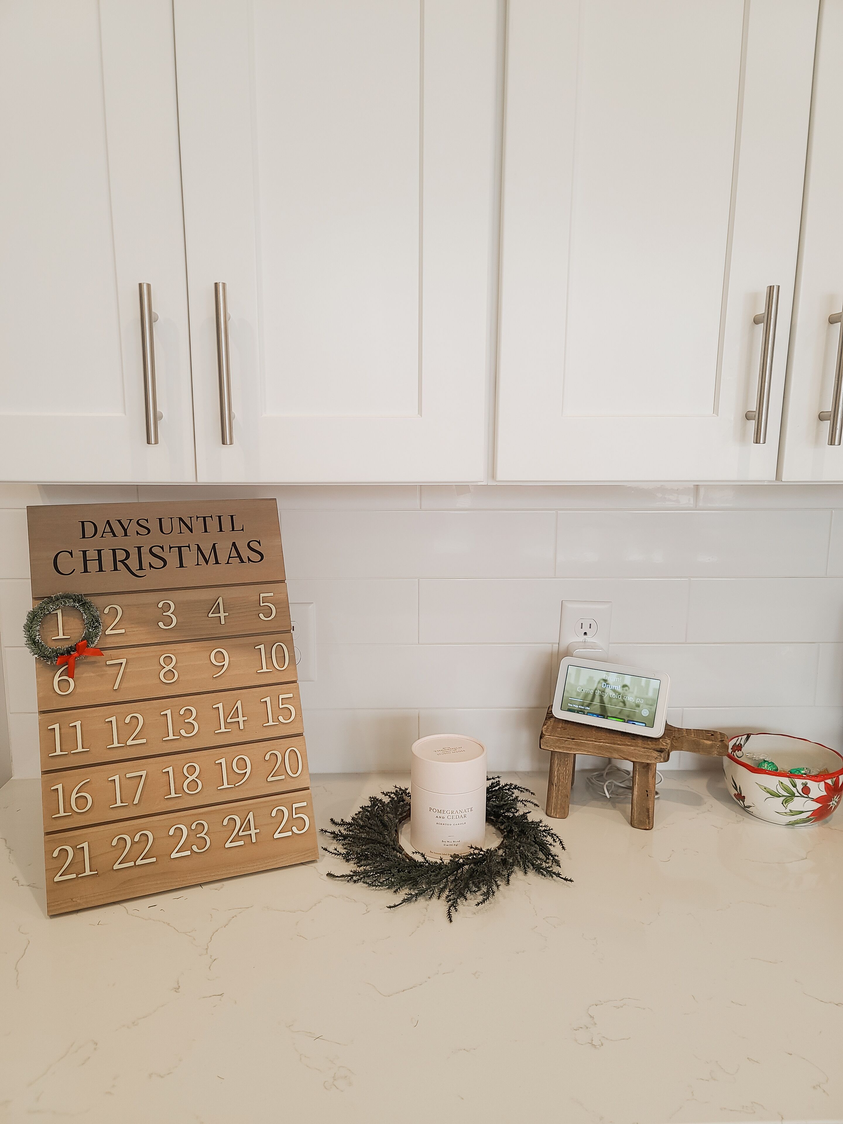 A kitchen with white cabinets, a wooden calendar, a small wooden stool, a candle, and a decorative wreath.