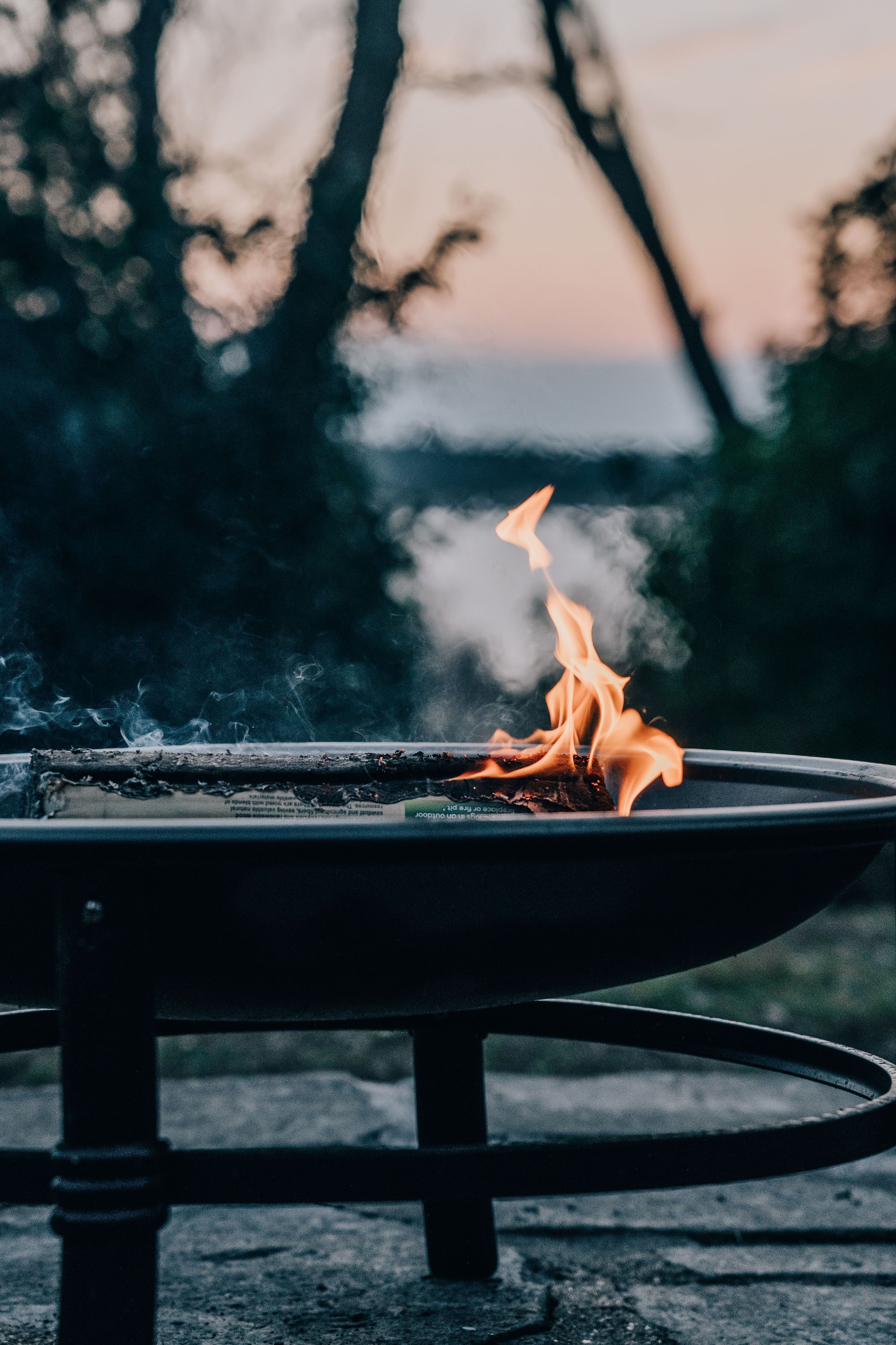 A campfire in a metal fire pit with a blurred background of trees and a sunset.