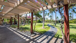 A covered outdoor area with a concrete pathway, palm trees, and a well-maintained lawn. - Upgraded Condo on Golf Course, Walking Distance to Beach and Shops! (Waikoloa)