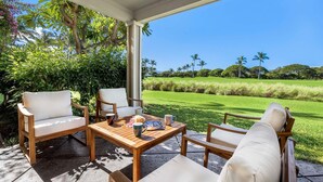 A covered outdoor seating area with wooden furniture and a table, overlooking a well-maintained lawn and distant trees. - Upgraded Condo on Golf Course, Walking Distance to Beach and Shops! (Waikoloa)