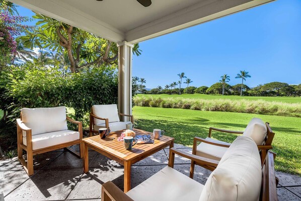 A covered outdoor seating area with wooden furniture and a table, overlooking a well-maintained lawn and distant trees. - Upgraded Condo on Golf Course, Walking Distance to Beach and Shops! (Waikoloa)