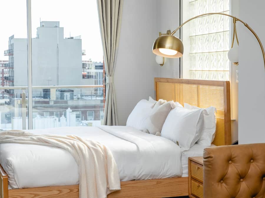 A bedroom with a large window, a bed with white bedding, a wooden headboard, and a gold floor lamp.