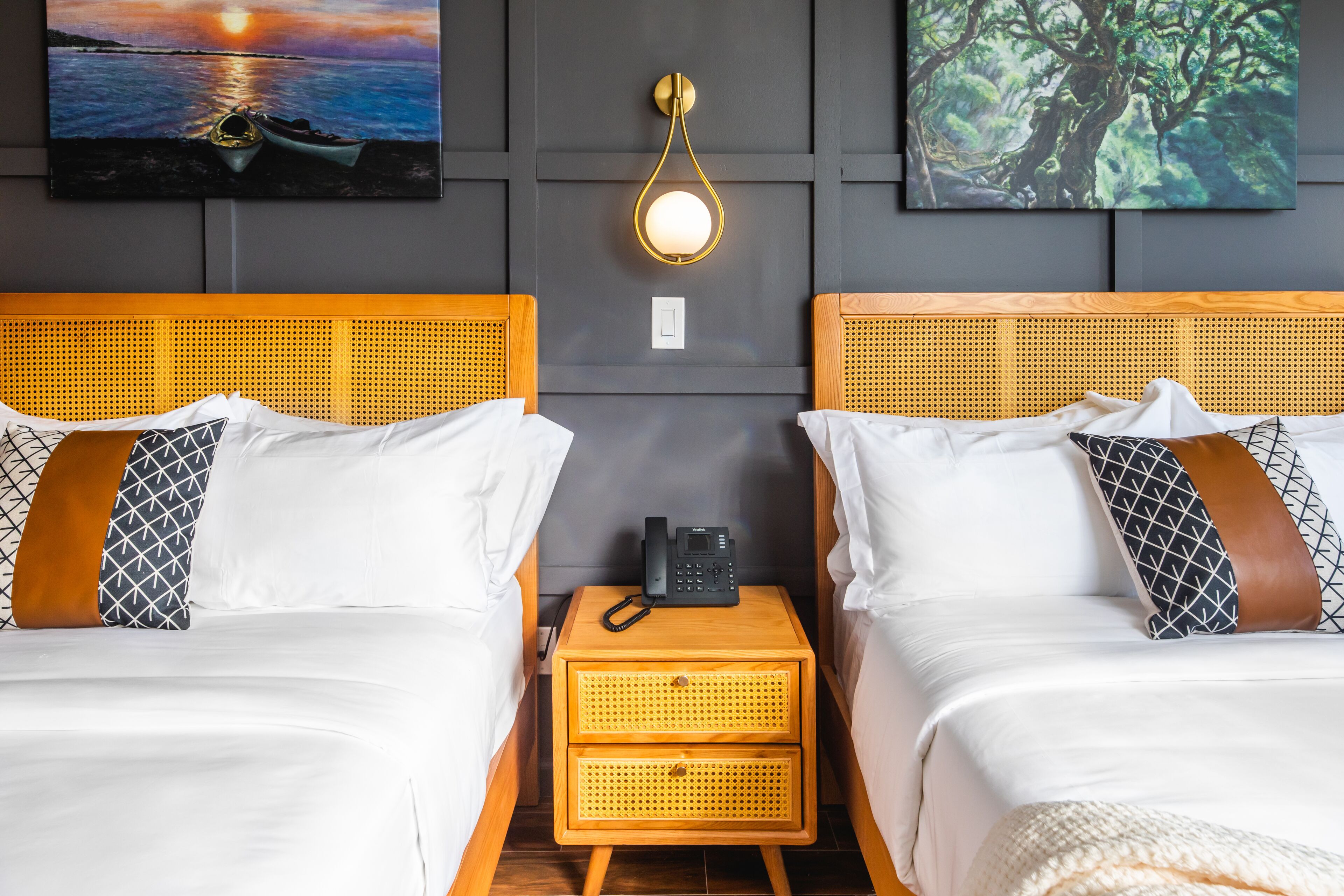 A hotel room with two beds, a nightstand with a phone, and wall art depicting a sunset and a forest.