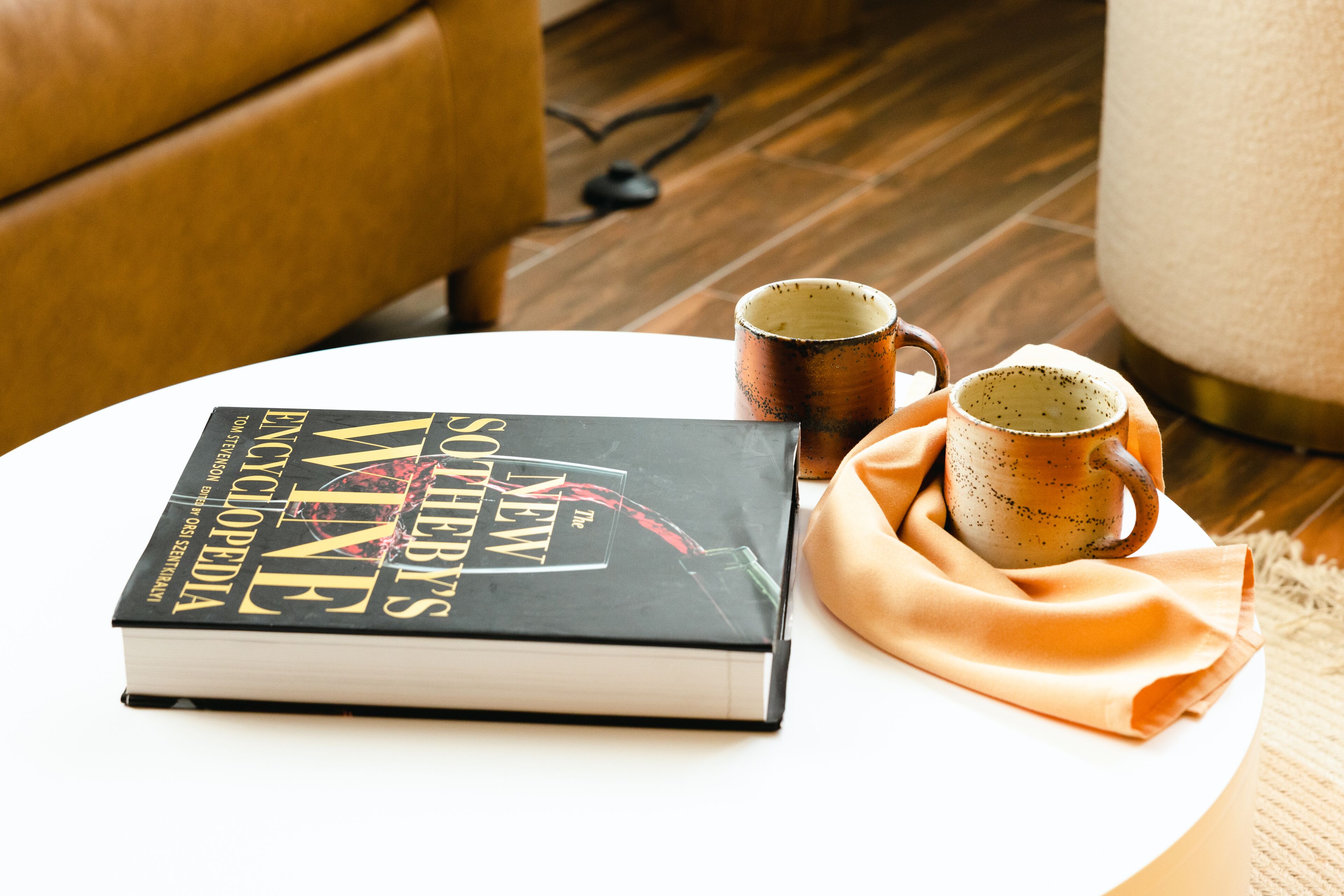 Un libro titulado "The New Wine Encyclopedia" sobre una mesa blanca, con dos tazas y un mantel de tela color beige.