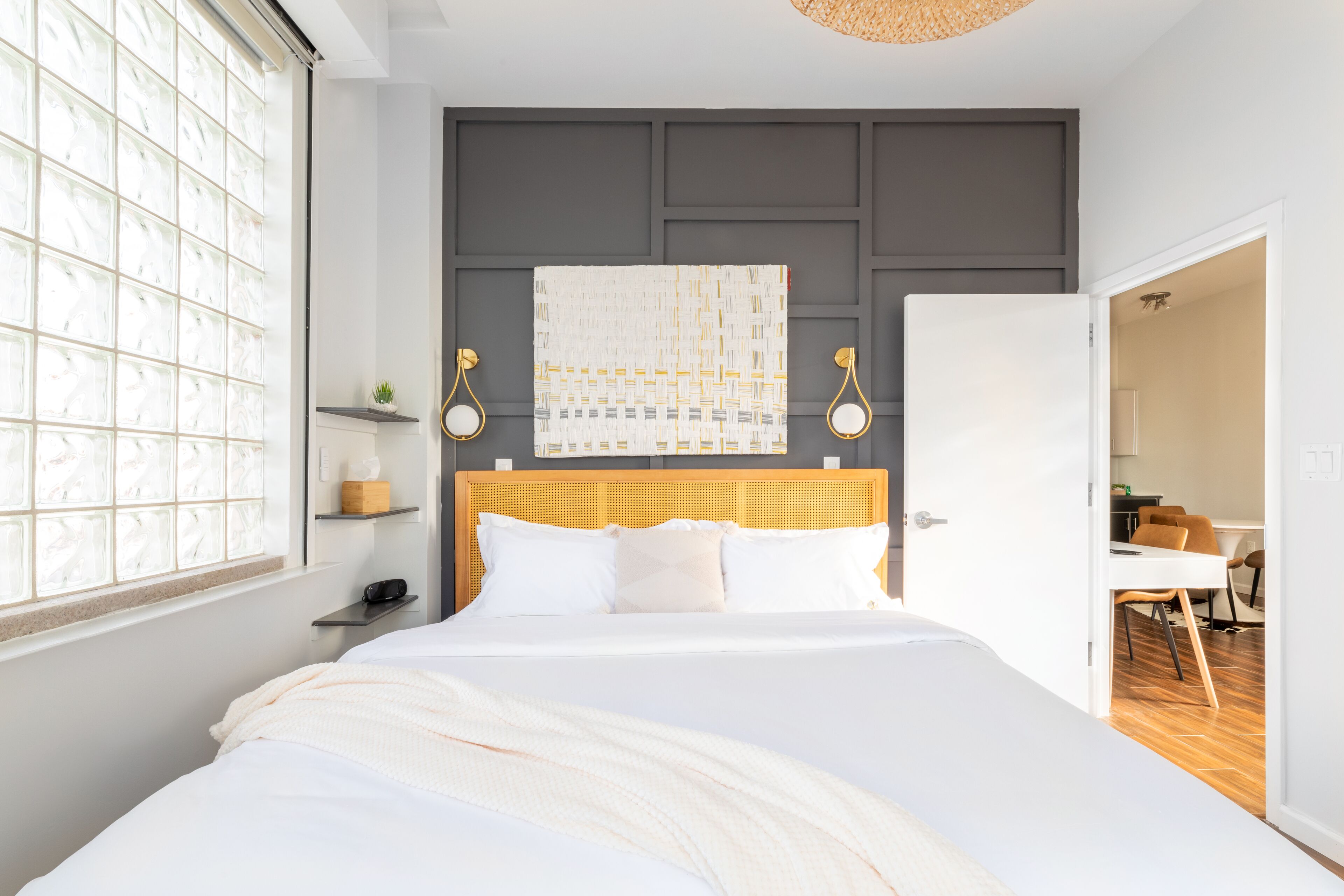 Quarto bem arrumado com uma cama grande, uma mesa de cabeceira com abajur, uma janela com vidro fosco e uma porta que dá para uma sala de jantar.