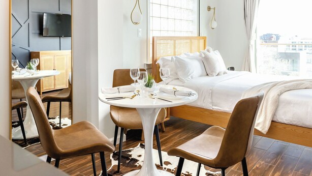 A hotel room with a bed, a dining table with chairs, a TV mounted on the wall, and a large window with curtains.