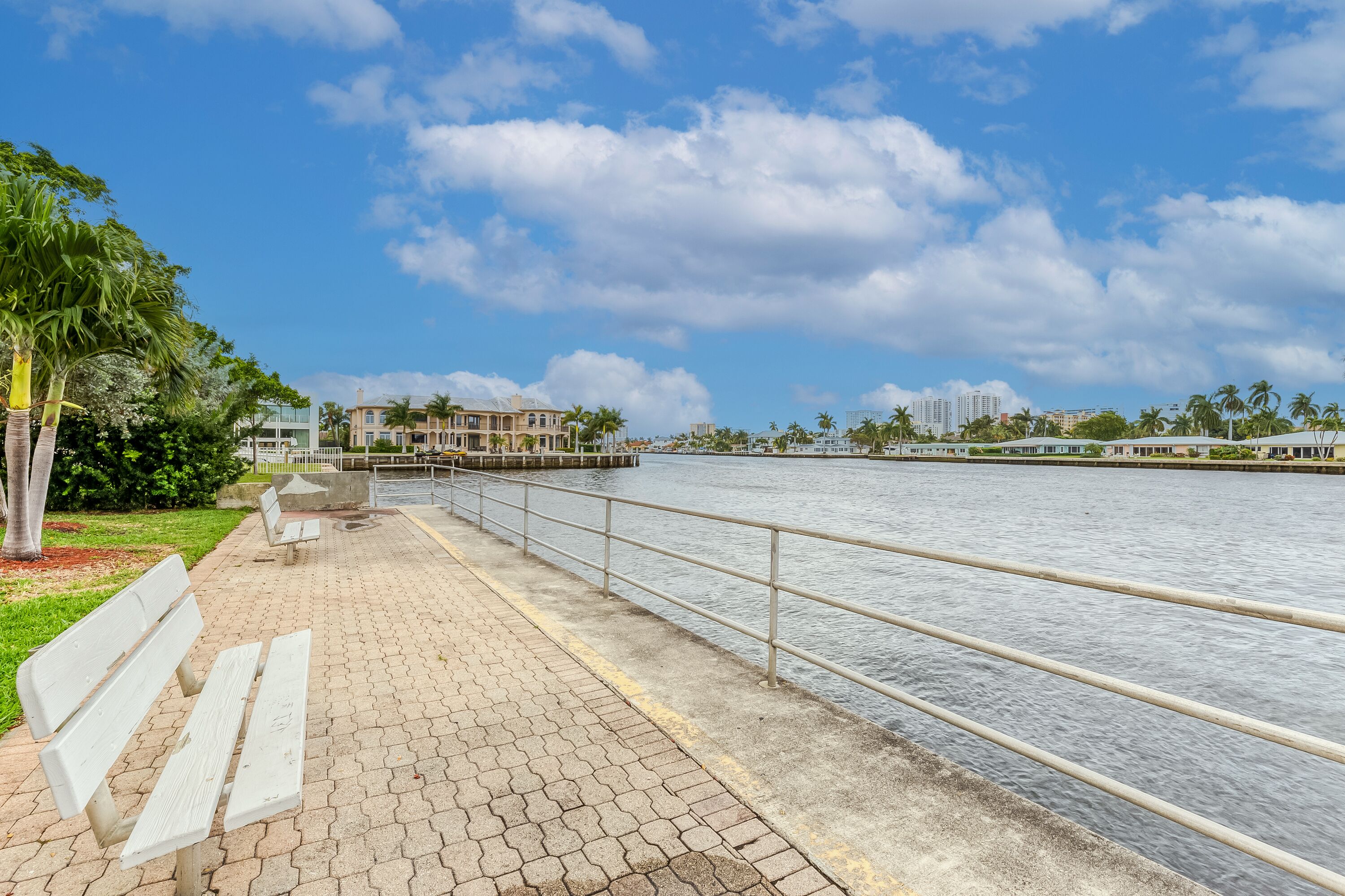 Un paseo marítimo con un banco, una barandilla metálica y vistas a un canal con edificios al fondo.