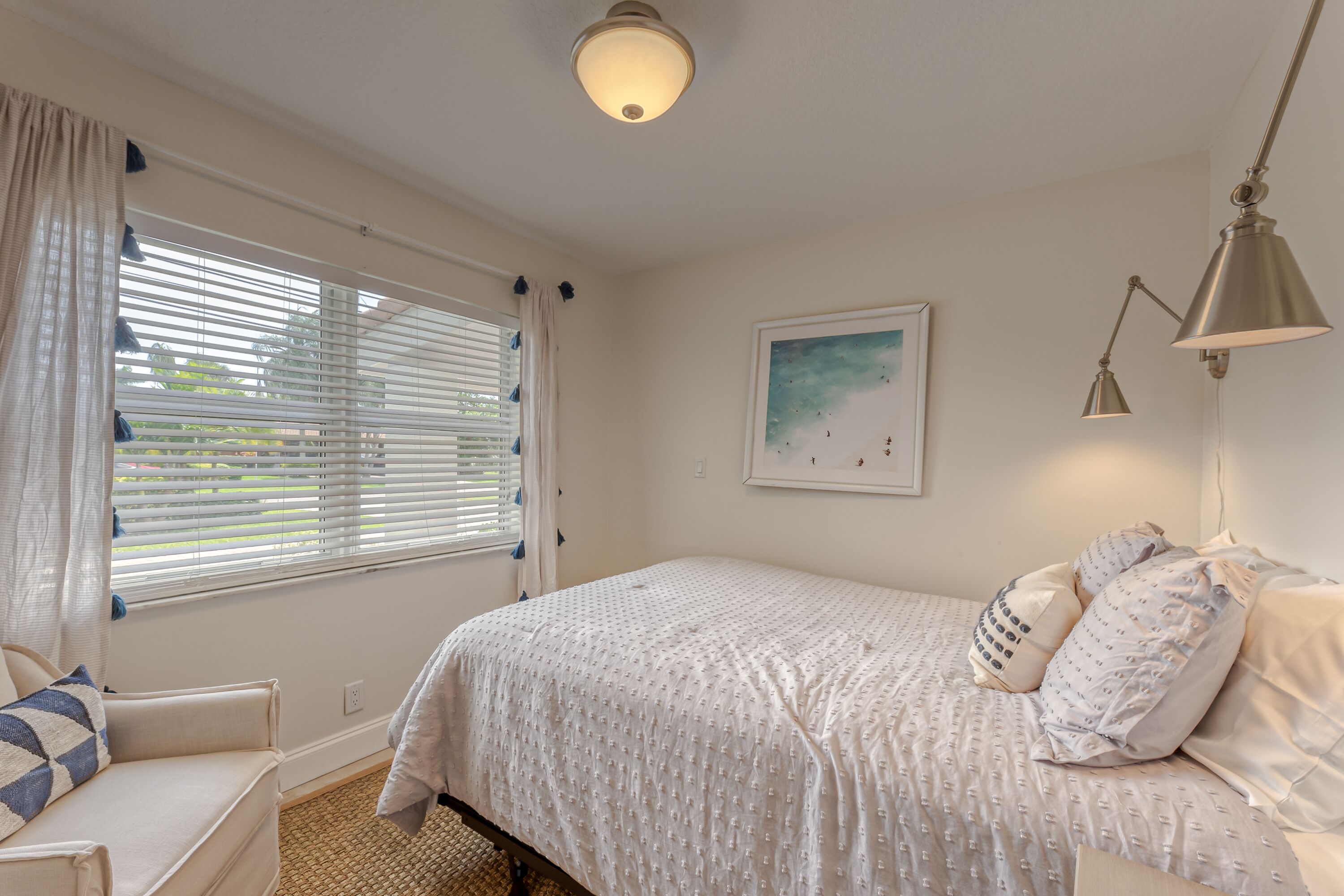 A bedroom with a bed, a chair, a window with blinds, a ceiling light, and a wall-mounted lamp.