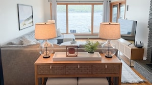 A modern living room with a large window overlooking a body of water, a sofa, a coffee table with decorative items, and a television.