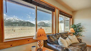Una habitación con sofá de cuero, dos lámparas, una mesa de madera y vistas a las montañas a través de amplias ventanas.
