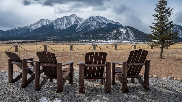 Tres sillas de madera tipo Adirondack mirando un paisaje montañoso con una valla y un árbol solitario.