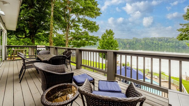 A deck with wicker furniture and a grill, overlooking a lake with a view of the surrounding greenery.