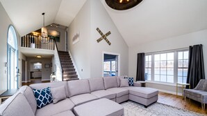 A spacious living room with a large sectional sofa, a staircase leading to an upper level, and a view of a lake through large windows.