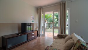 A living room with a TV on a wooden cabinet, a sofa with patterned cushions, and a sliding glass door leading to a garden. - Studio Garden, cozy and unique (SC)