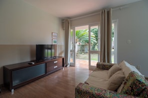 A living room with a TV on a wooden cabinet, a sofa with patterned cushions, and a sliding glass door leading to a garden.