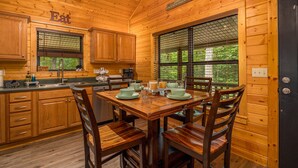 Interior de una cabaña de madera con una mesa de comedor para cuatro personas, armarios de madera y una ventana que ofrece vistas a la vegetación.