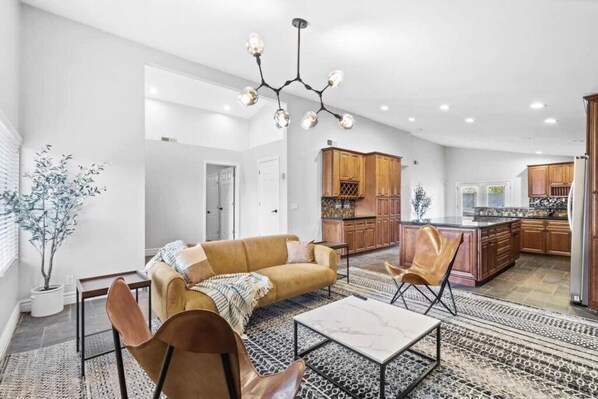 A spacious living area with a sofa, chairs, and a coffee table. There is a kitchen in the background with wooden cabinets and a central island.