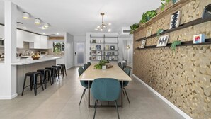 Una cocina moderna con una isla central, un comedor con una mesa larga y sillas, y una pared decorativa con patrones geométricos y estanterías.