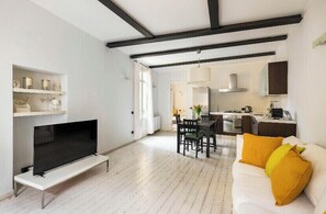 Un salon moderne avec un téléviseur à écran plat, un coin repas avec une table et des chaises, un canapé avec des coussins jaunes et une cuisine en arrière-plan.