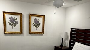A bedroom with a wooden bed, white bedding, two framed botanical prints on the wall, a nightstand with a lamp, and a ceiling fan.
