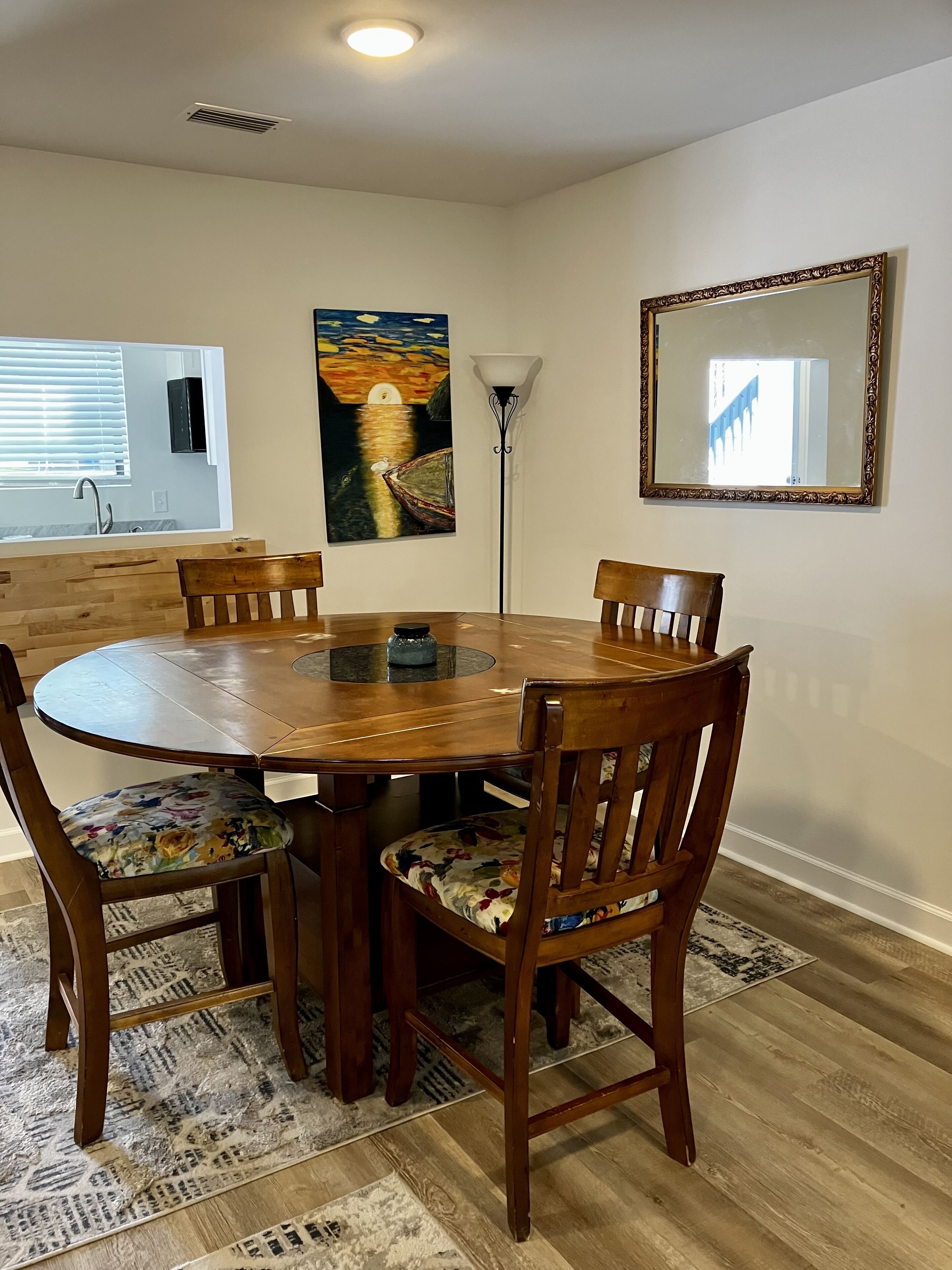 Une salle à manger avec une table ronde en bois, quatre chaises, un tableau, un lampadaire et un miroir au mur.