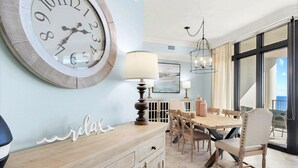 Un coin repas comprenant une grande horloge murale, une table avec des chaises, une lampe et une vue sur l’extérieur.