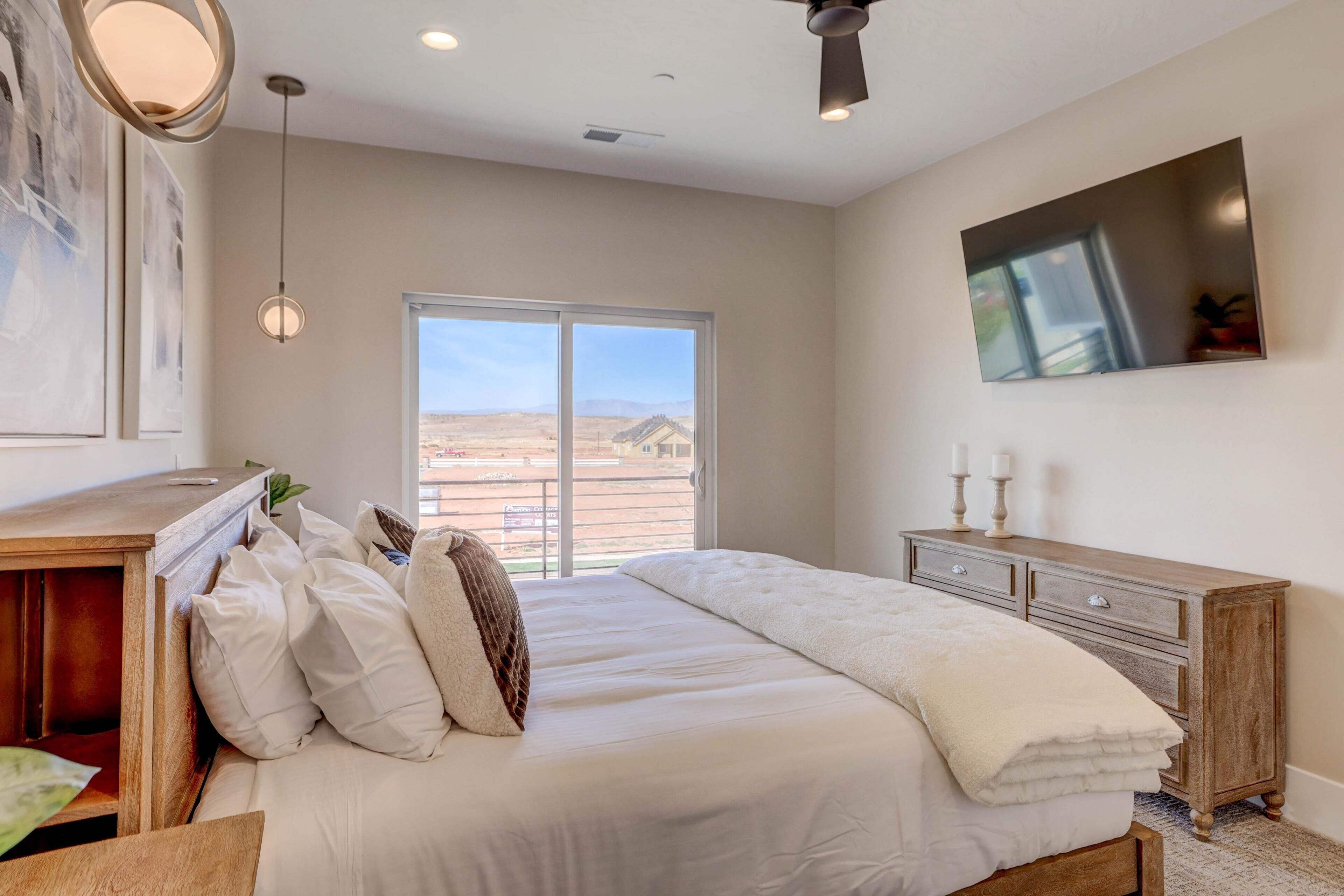 A bedroom with a large bed, a wooden dresser, a ceiling fan, and a large window with a view of a desert landscape.