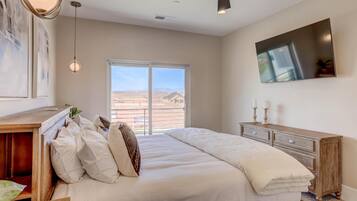 A bedroom with a large bed, a wooden dresser, a ceiling fan, and a large window with a view of a desert landscape.