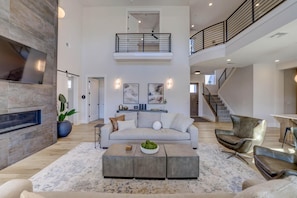 A spacious living area with a high ceiling, a fireplace, a sofa, a coffee table, and a staircase leading to an upper level.